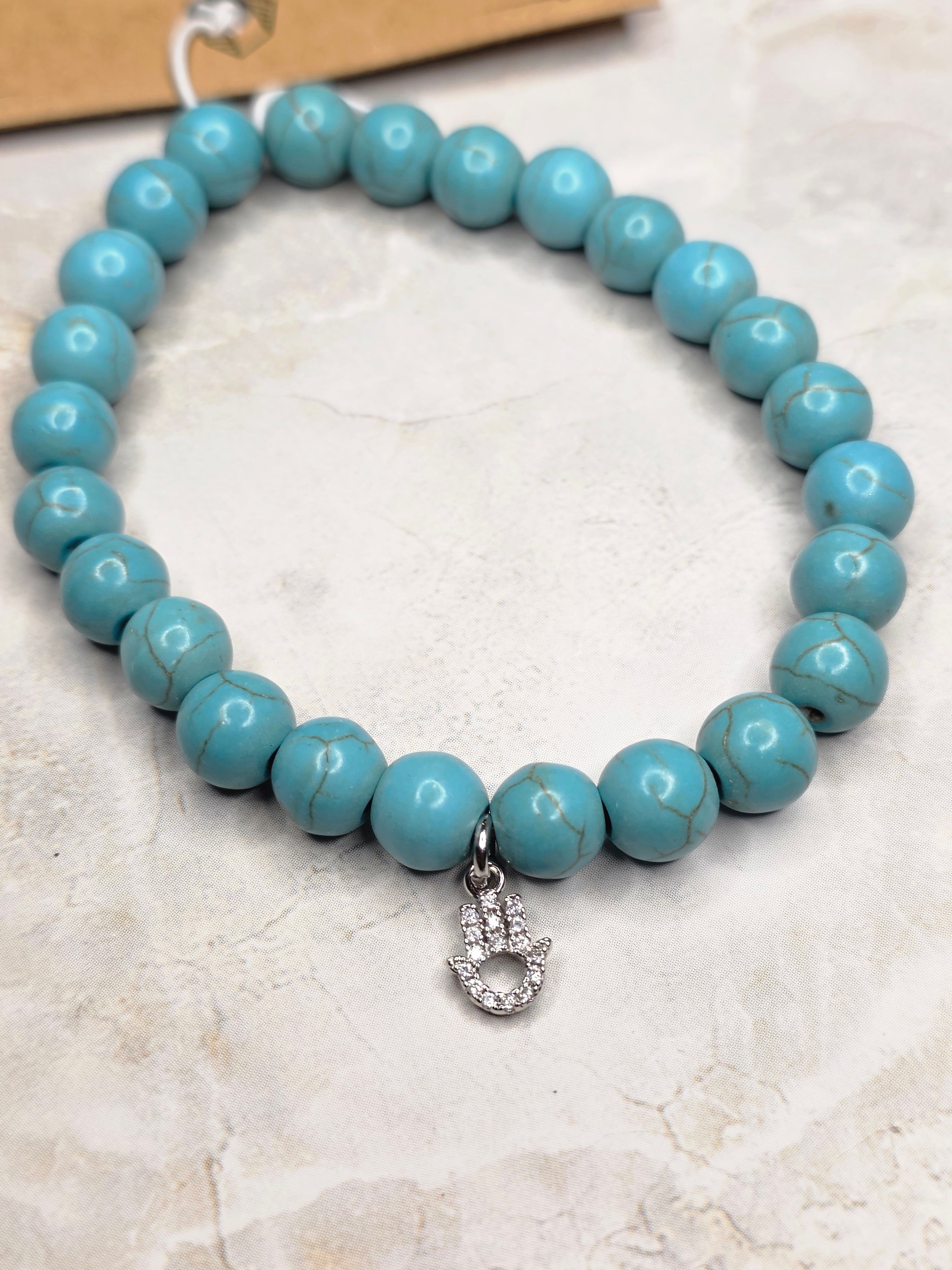 A turquoise beaded bracelet with a silver-toned crescent moon charm, displayed on a textured beige stone surface.
