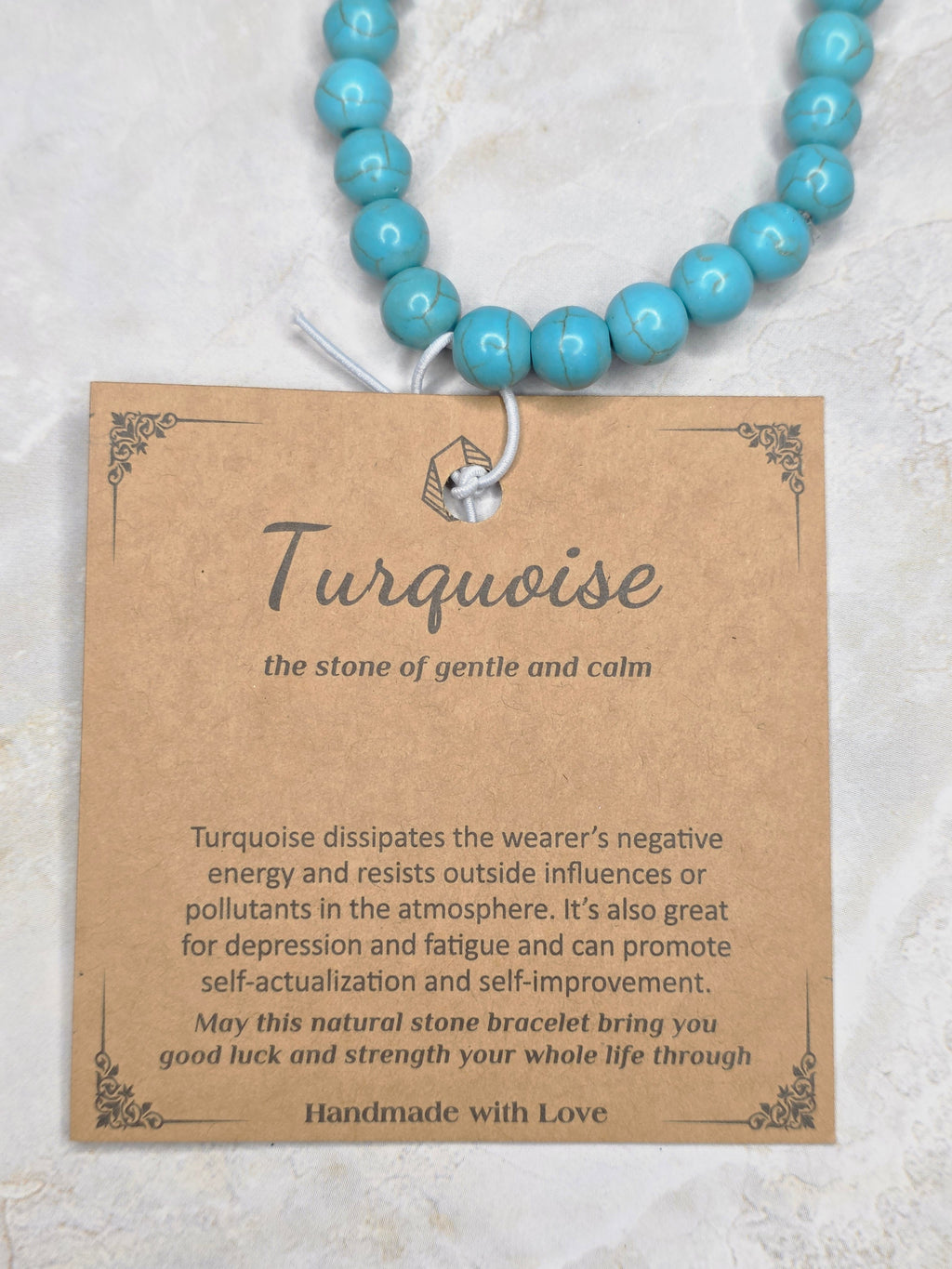 A turquoise beaded bracelet with a white cord and a brown kraft paper tag featuring spiritual text about turquoise, laid out on a light textured background.