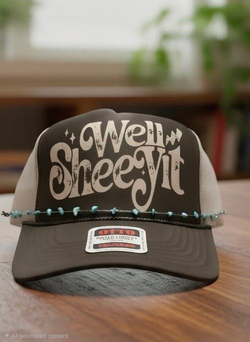 A dark brown and beige trucker hat with 'Well Sheyit' written on the front, resting on a wooden table with a blurred indoor background.