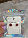A pair of round stud earrings with deep red glittery centers and gold-toned edges, displayed on a white card inside a holographic zip pouch and held by a hand with purple striped nail polish.