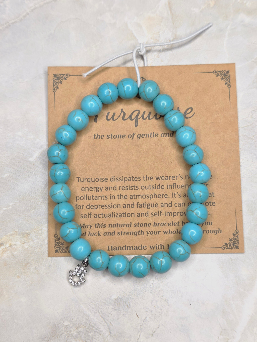 A turquoise beaded bracelet with a silver-toned charm, displayed on a brown kraft paper card with printed text, set against a light beige stone background.