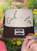 A personalized trucker hat with a cream front panel and dark brown mesh back, held by a hand. The front panel features the custom name 'Gah - Lee' in black script, decorated with a pearl beaded trim across the brim.