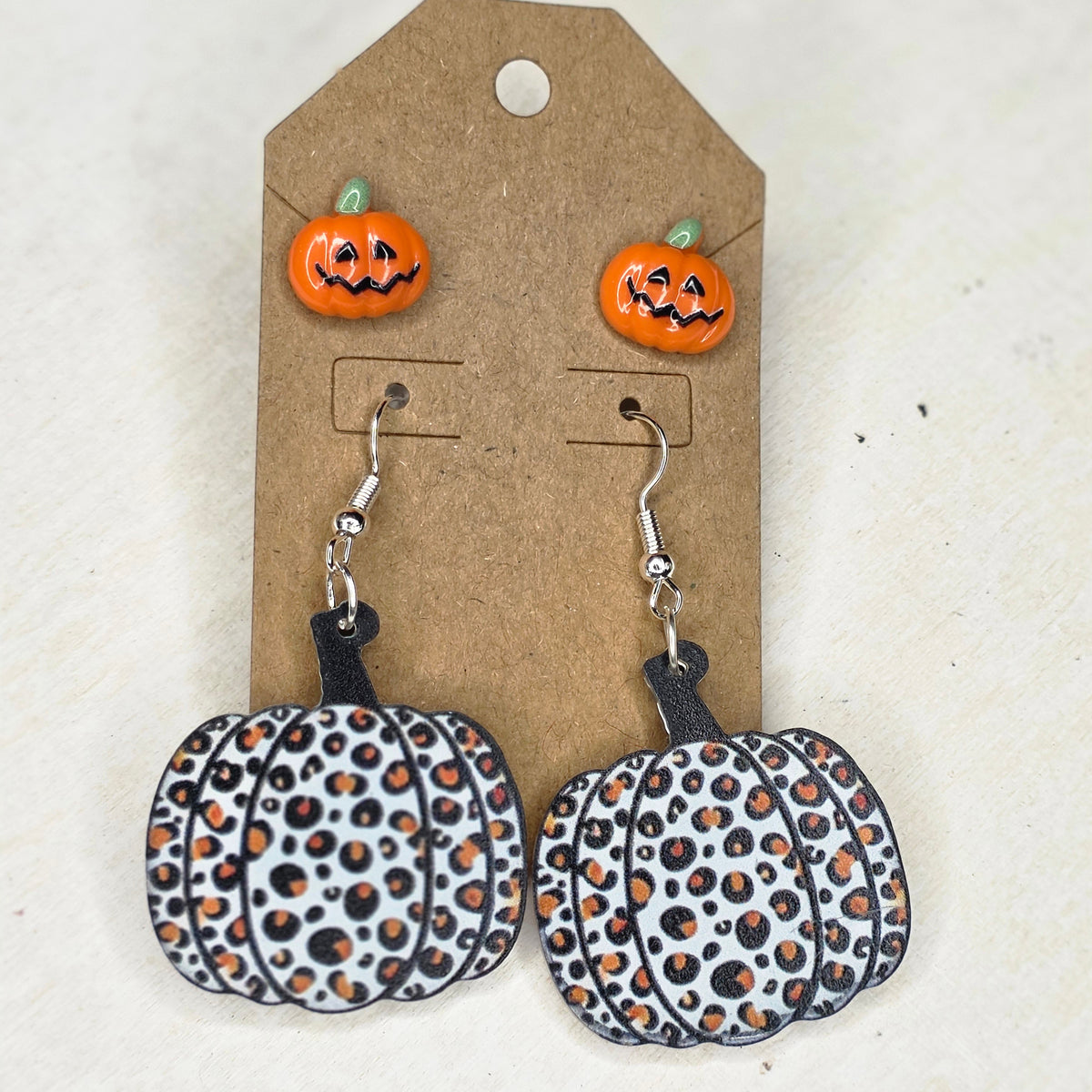 Leopard-print pumpkin dangle earrings and orange jack-o'-lantern stud earrings on brown display card