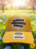 A mustard yellow trucker cap with a floral graphic and the text 'WILD FLOWERS DON'T CARE WHERE THEY GROW' on the front, displayed on a grassy field with flowers and blurred trees in the background.