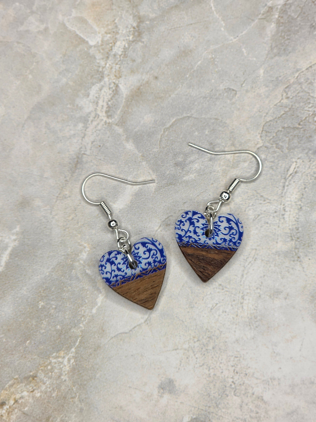 A pair of handmade heart-shaped dangle earrings featuring a blue and white patterned top half and a dark brown wood grain bottom half, suspended from silver fishhook wires, displayed on a light beige stone surface.