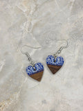 A pair of handmade heart-shaped dangle earrings featuring a blue and white patterned top half and a dark brown wood grain bottom half, suspended from silver fishhook wires, displayed on a light beige stone surface.